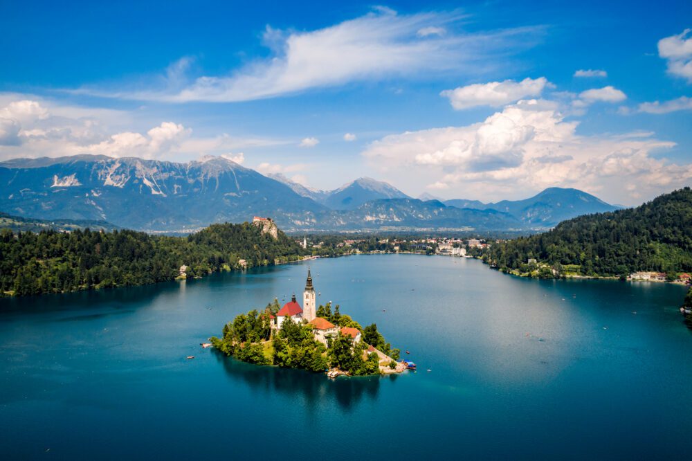 Aerial view of the church spire on forested Bled Island in sapphire-blue Lake Bled with the Julian Alps behind in Slovenia